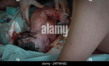 Immagine di nascita autentica: Una bambina appena nata piangente con i capelli scuri coperti di sangue pochi istanti dopo la nascita. Cordone ombelicale ancora attaccato. Foto Stock