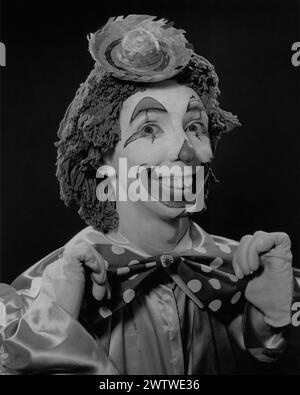 Primo piano di un uomo con un vestito da clown, volto dipinto, grande cravatta, testa per mocio e un cappello di paglia mini sulla testa Foto Stock