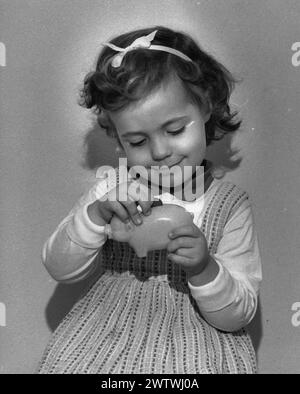 Bambina in abito con un fiocco tra i capelli, che mette una moneta nel salvadanaio Foto Stock