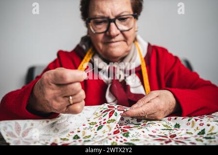 primo piano ritratto di una donna anziana vestita di rosso, sarta, cucita a mano un indumento di stoffa nel soggiorno bianco Foto Stock