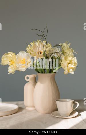 Mazzo di gerbera gialla, tulipani e ranunculus in un vaso con ceramiche assortite su un tavolo alla luce del sole Foto Stock