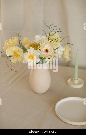 Mazzo di fiori gialli di gerbera, tulipani e ranuncolo in un vaso su un tavolo con candela e piatto Foto Stock