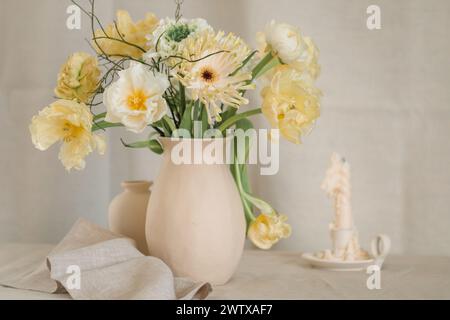 Mazzo di fiori gialli di gerbera, tulipani e ranunculus in un vaso su un tavolo con una candela e un tovagliolo Foto Stock