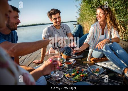 Gruppo di amici che si diverte a fare un picnic vicino a un lago, seduti sul molo a mangiare e bere vino. Foto Stock