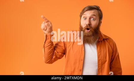 Uomo barbuto stupito che punta allo spazio vuoto su sfondo arancione Foto Stock
