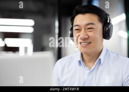 Uomo d'affari asiatico professionista con una camicia blu, felice e sorridente con le cuffie indossate, che lavora nel suo ufficio. Foto Stock