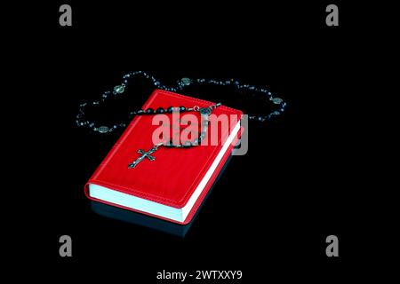 Nuovo Testamento con crocifisso rosario e perline su una superficie nera riflettente Foto Stock