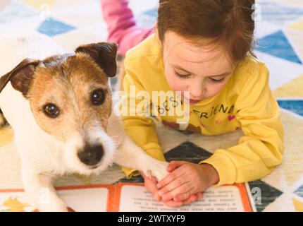 Piccola ragazza che impara a leggere. La bambina sta leggendo un libro al suo cane da compagnia. Foto Stock