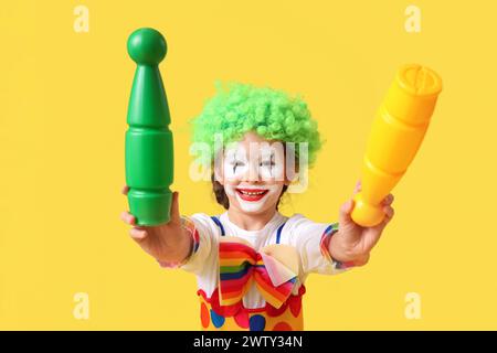 Divertente bambina in costume da clown con mazze da giocoliere su sfondo giallo. Festa del Fool's Day di aprile Foto Stock