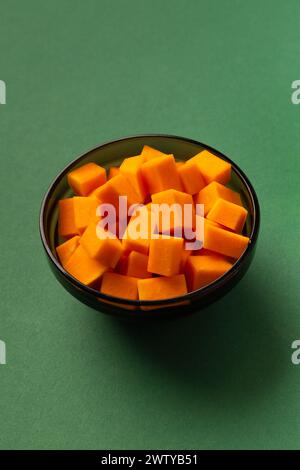 Zucca dolce di colore arancio tagliata a cubetti in un piatto nero su sfondo verde. Modello culinario per piatti di zucca. Mangiare sano. Fette di zucca. Foto Stock