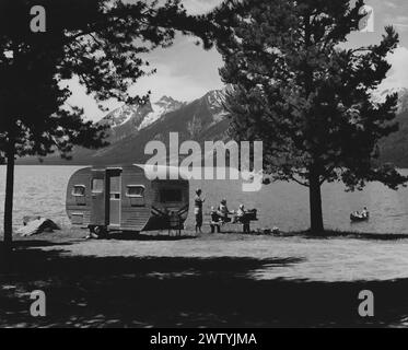 Madre e due bambini seduti a un tavolo da picnic accanto al loro rimorchio che è seduto sulla riva di un lago, una catena montuosa sullo sfondo. Padre e un altro bambino sono in canoa sul lago Foto Stock