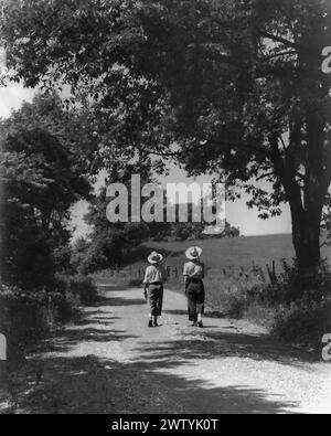 Due persone camminano lungo una strada sterrata, indossando cappelli di paglia e trasportando pali da pesca, allontanandosi dalla telecamera mentre passano sotto un grande albero ombreggiato Foto Stock