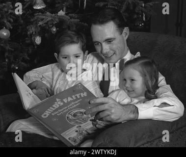 Il giovane papà con una camicia e una cravatta seduto su una poltrona di fronte all'albero di Natale con i suoi due bambini in pigiama seduti in grembo mentre leggono un libro insieme Foto Stock