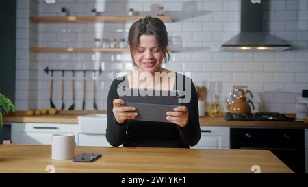 Donna che gioca a un videogame su un tablet mentre è seduta in cucina a casa. Donna eccitata e concentrata, coinvolta in un interessante gioco di corse Foto Stock