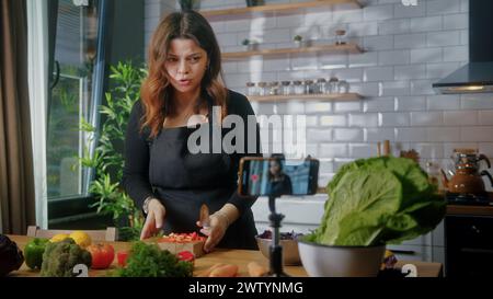Una ragazza carina in grembiule in piedi in cucina registra su smartphone il nuovo videoblog di cibo. Donna che taglia verdure su un tagliere, blogging, lifest sano Foto Stock