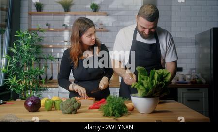 Coppia che ride, si diverte a preparare un sano pasto vegetariano con insalata per una cena romantica nella cucina domestica di casa Foto Stock