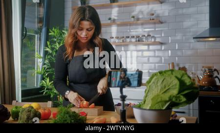 La donna in grembiule in piedi in cucina registra su smartphone il nuovo videoblog alimentare. Donna che taglia verdure su un tagliere, blogging, stile di vita sano, Foto Stock