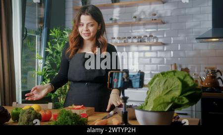 La giovane donna in grembiule in piedi in cucina registra su smartphone il nuovo videoblog alimentare. Donna che taglia verdure su un tagliere, blogging, sano l Foto Stock