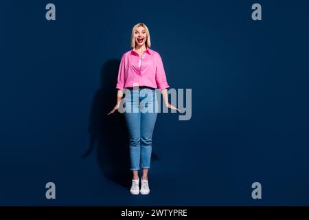 Foto a grandezza naturale di jeans con camicia rosa, una donna entusiasta e allegra, in piedi sulle punte delle dita come una bambola isolata su sfondo blu scuro Foto Stock