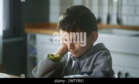 Bambino turbato seduto in cucina da solo a casa. Uno scolaro solitario che si sente deluso, vittima di bullismo e stressato seduto a tavola. Problemi con i bambini delle scuole Foto Stock