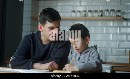 Papà aiuta lo scolaro con i compiti a casa. Papà aiuta il figlio a scrivere mentre è seduto in cucina. Genitori adulti o tutor con bambini che imparano. Foto Stock