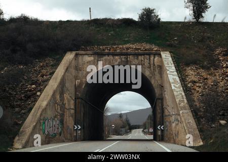 Strada asfaltata vuota che passa attraverso un tunnel a forma di arco. Fotografia di viaggio scura e drammatica. Viaggia in un paesaggio buio e freddo con nuvole piovose Foto Stock