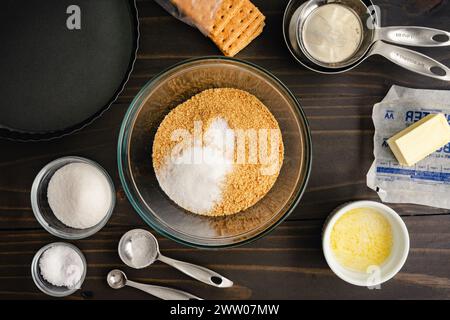 Ingredienti della crosta Graham Cracker con una padella: Biscotti sbriciolati Graham e zucchero in una ciotola da miscelare circondata da altri ingredienti e utensili da cucina Foto Stock