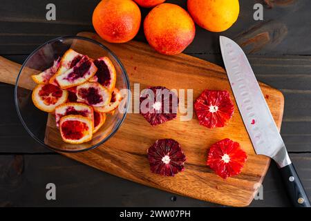 Arance sanguigne sbucciate e deseminate su un tagliere di legno: Arance pelate, tagliate a metà e intere di lamponi con un coltello dello chef e bucce laterali Foto Stock