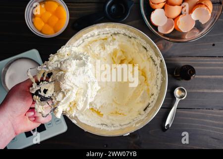 Mano che tiene in mano una frusta per il mixer con supporto ricoperta di crema Buttercream: Mano della donna che tiene una frusta metallica ricoperta di glassa di crema di burro di meringa svizzera Foto Stock