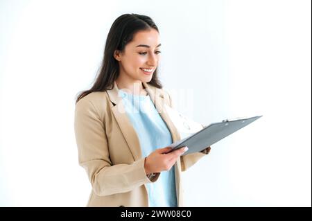 Bella e bella donna d'affari indiana o araba, consulente finanziario, segretaria, si erge su sfondo bianco isolato, tenendo una cartella con documenti e grafici, guardandola, sorridendo Foto Stock