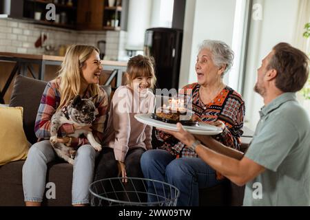 Una scena emozionante si svolge mentre una famiglia multigenerazionale si riunisce su un divano per presentare una torta di compleanno a una nonna adorata, creando ricordi indimenticabili Foto Stock