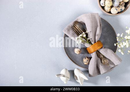 Tovagliolo con posate vintage e uova di quaglia su piatto grigio, fiori bianchi, coniglietti in ceramica. Vista dall'alto. Elegante tavolino pasquale grigio per caffè e ristoranti Foto Stock