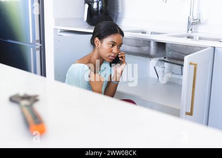 Giovane afroamericano accovacciato da un lavandino rotto in cucina che fa una telefonata Foto Stock