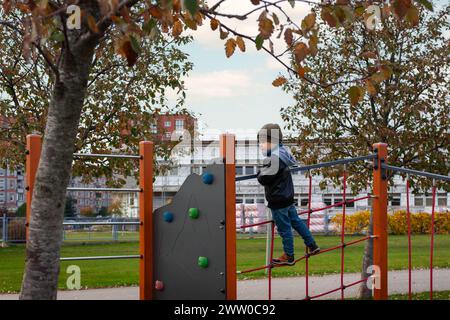 In autunno un bambino carino gioca a sport nel parco giochi per bambini in giardino. Il bambino sale coraggiosamente la scala di corda. Foto Stock