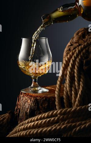 Il brandy viene versato da una bottiglia in un bicchiere. Bicchiere con whisky, cognac o brandy su un vecchio ceppo. Foto Stock