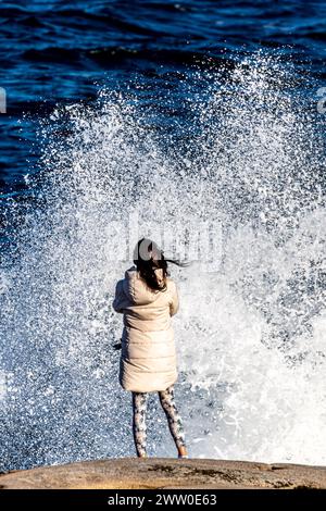 Donna in piedi sulla scogliera con la schiena rivolta verso la telecamera. Schiuma bianca visibile dall'onda che si schianta. Sfondo marino nell'immagine monocromatica Foto Stock