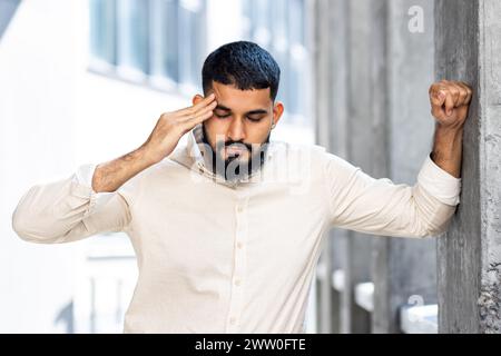 Foto ravvicinata di un giovane arabo con una camicia e jeans in piedi fuori con gli occhi chiusi, appoggiato contro il muro di un edificio e tenendo la testa. Prova forte pressione e dolore. Foto Stock