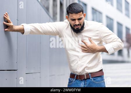 Un maschio adulto viene visto fuori, sembra angosciato mentre stringe il petto nel dolore. La sua espressione e postura suggeriscono che sta vivendo un attacco di cuore o un problema medico acuto, sottolineando l'importanza della consapevolezza della salute. Foto Stock