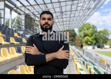 Un giovane uomo barbuto in abbigliamento sportivo si erge con fiducia con le braccia incrociate in uno stadio vuoto, trasudando determinazione. Foto Stock