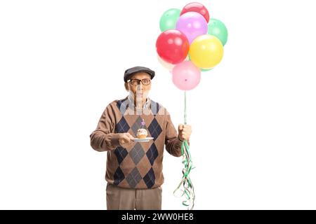 Uomo anziano che regge un cupcake di compleanno con una candela e un mucchio di palloncini isolati su sfondo bianco Foto Stock