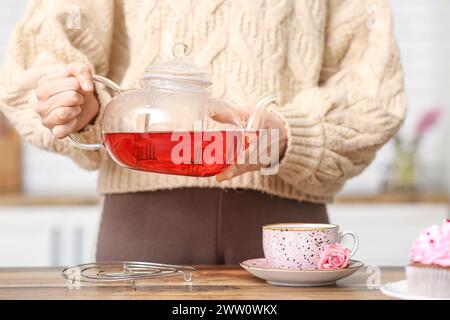 Giovane donna che versa tè all'ibisco dalla teiera nella tazza in cucina Foto Stock