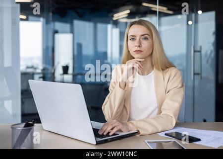 Donna d'affari professionale con capelli biondi, concentrata sul lavoro alla scrivania dell'ufficio con un computer portatile, trasudando fiducia e determinazione. Foto Stock