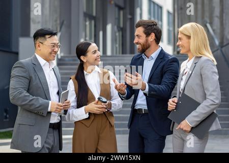 Quattro professionisti sorridenti che organizzano riunioni informali all'aperto, con tablet e smartphone in un ambiente urbano moderno. Foto Stock