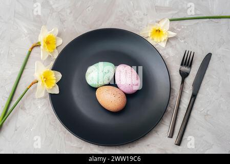 Piatto nero con uova di Pasqua, fiori di narcidi gialli e posate su sfondo grigio testurizzato. Vista dall'alto, disposizione piatta. Foto Stock