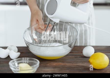 La mano della donna sta sbattendo le uova bianche con il mixer elettrico nel recipiente di vetro, la ricetta del forno, lo sfondo della cucina Foto Stock
