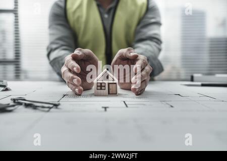 Le mani dell'ingegnere architetto tengono e proteggono il mini modello di casa sul progetto. Protect House, progettazione di pianificazione dei costi e concetto di costruzione. Foto Stock