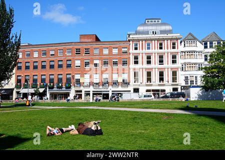 Negozi e caffetterie sul marciapiede lungo Cathedral Yard con giovani donne sdraiate sull'erba in primo piano, Exeter, Devon, Regno Unito, Europa. Foto Stock