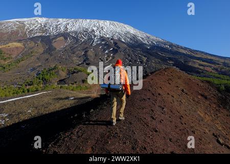 L'escursionista con lo zaino sul retro cammina lungo il crinale del cratere vulcanico estinto nel Parco dell'Etna, sullo sfondo innevato e ripido pendio del sud-ovest Foto Stock