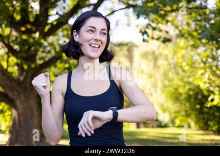 Foto ravvicinata di una giovane donna felice che fa sport e fa jogging nel parco, guardando un orologio intelligente e mostrando un gesto di successo con la mano, gioendo della vittoria. Foto Stock