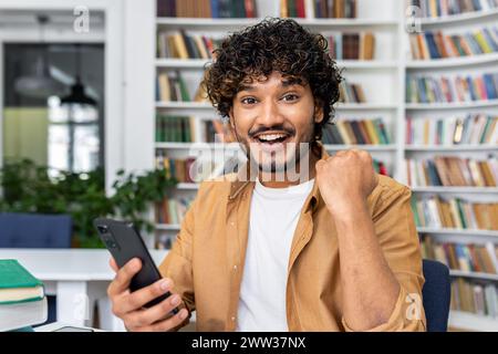 Gioioso giovane adulto in una camicia casual che celebra il successo mentre tiene in mano un telefono cellulare, con sfondo da libreria. Foto Stock